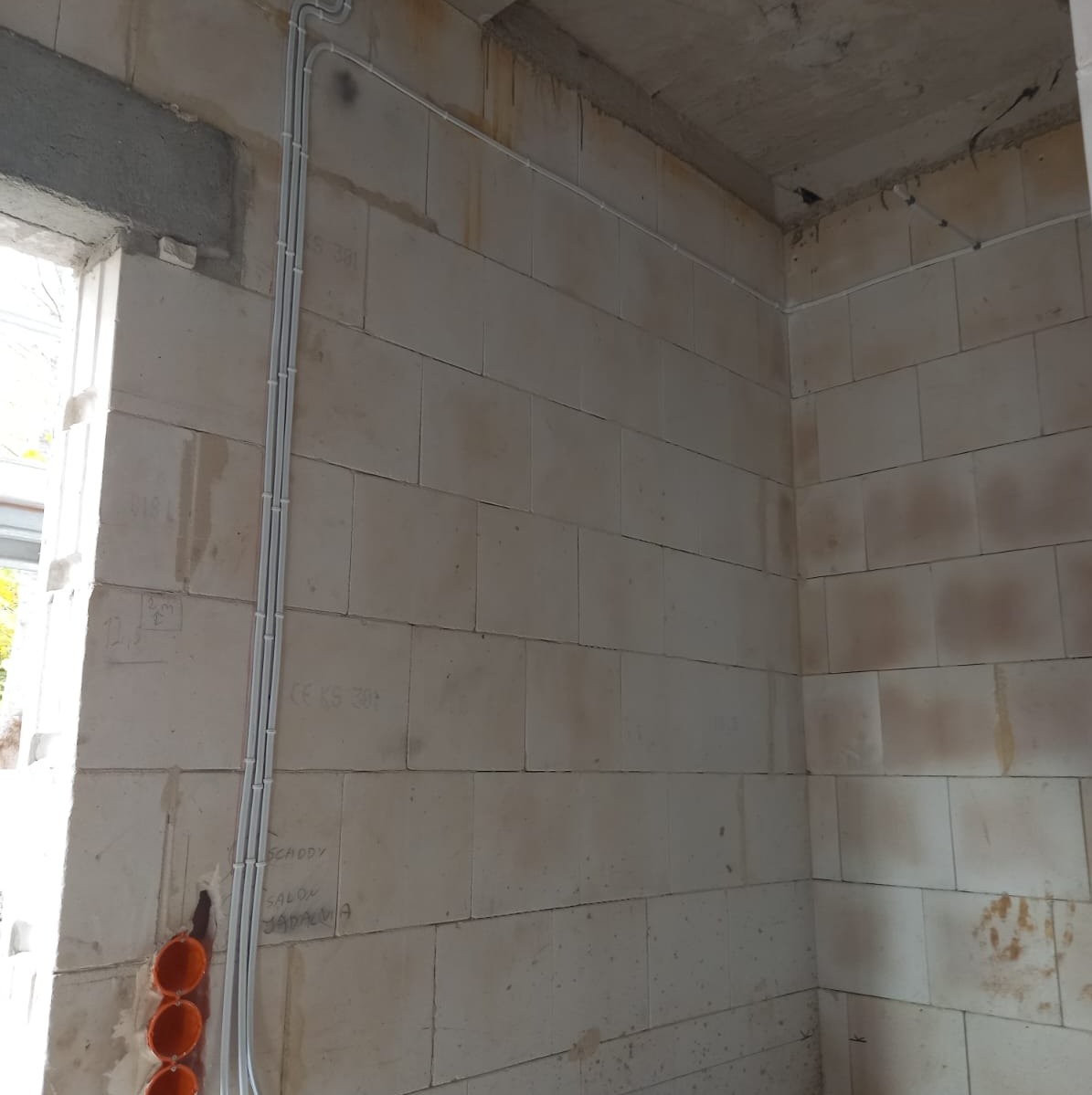 Wewnętrzna instalacja elektryczna w budynku w stanie surowym, widoczne kable ułożone w peszlach na ścianie z betonu komórkowego, puszki instalacyjne w kolorze pomarańczowym.