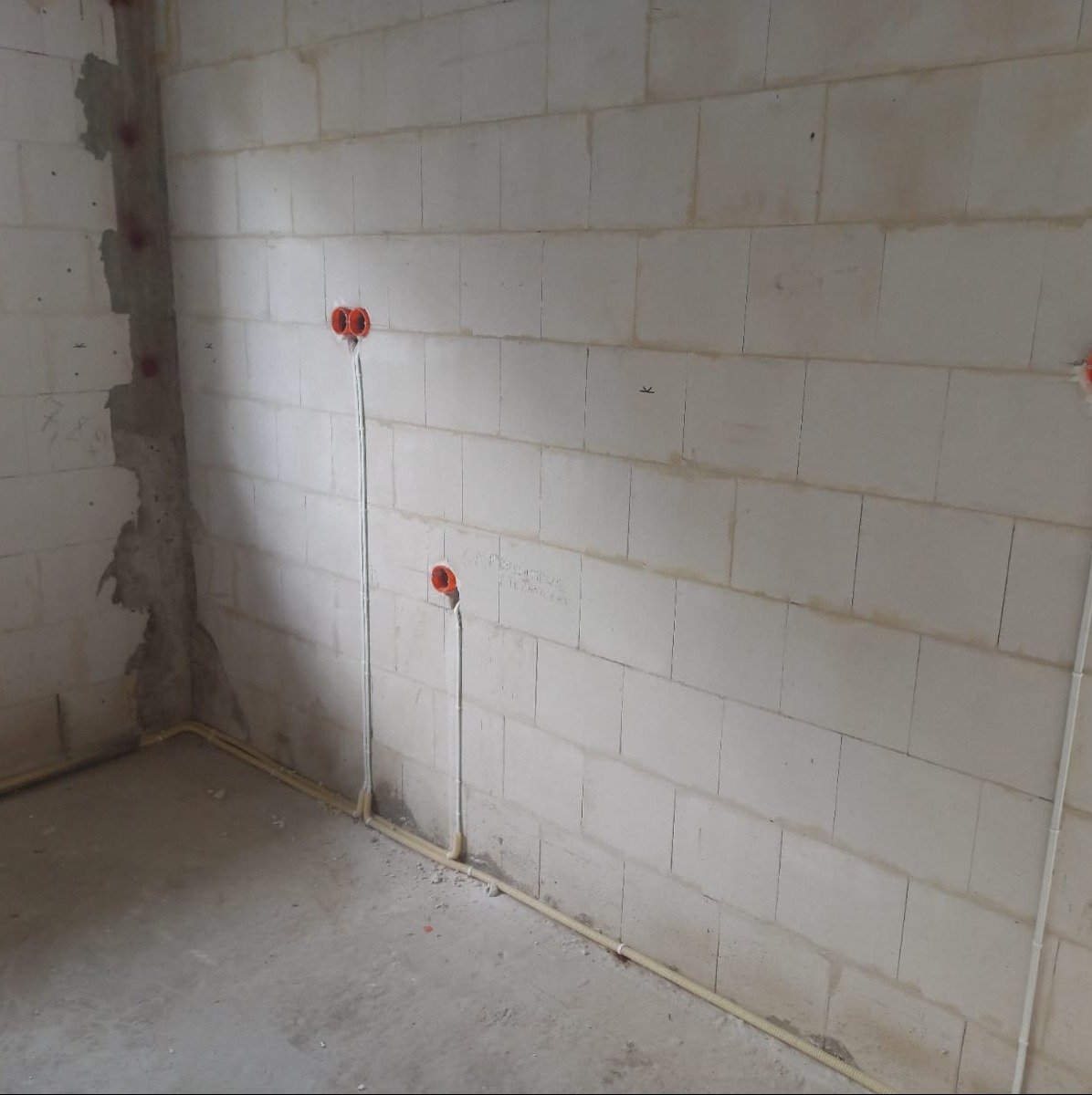 Surowy stan: Ściana z betonu komórkowego z widocznymi pomarańczowymi puszkami instalacyjnymi i białymi przewodami elektrycznymi, częściowo ukrytymi w żółtych rurkach ochronnych, na podłodze szary...