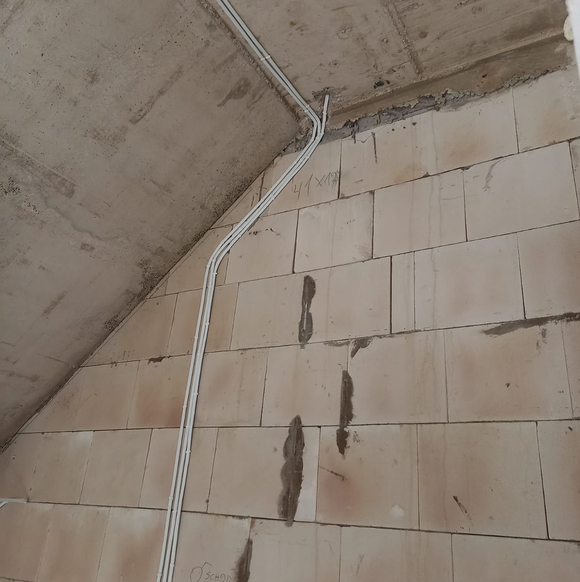Białe rurki instalacyjne prowadzone po surowej ścianie z bloczków, widoczne ślady po zaprawie i notatki odręczne na ścianie.