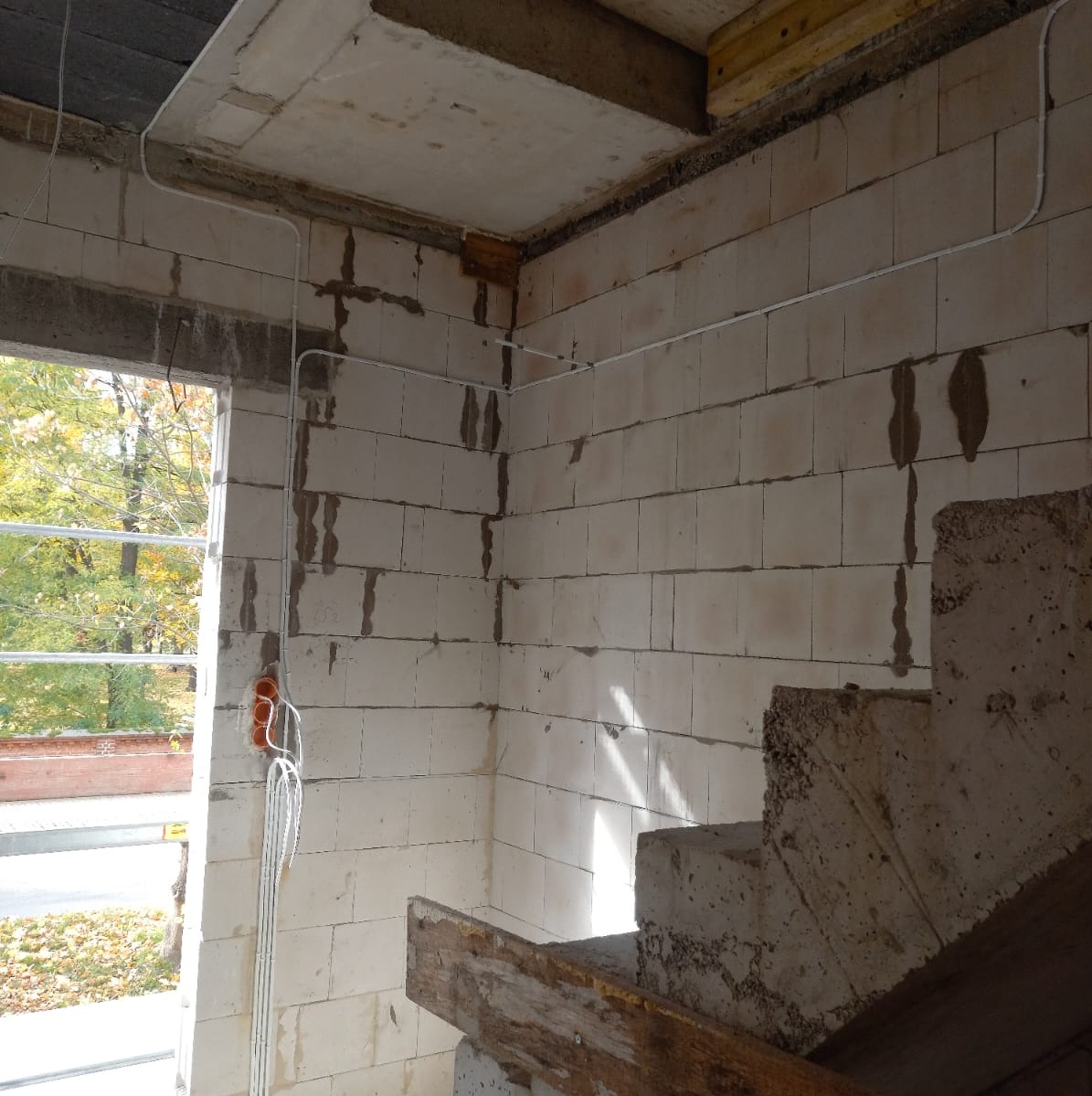 Surowe wnętrze w trakcie budowy z widocznymi przewodami elektrycznymi poprowadzonymi po ścianach z białego bloczka, pomarańczowa puszka instalacyjna przy oknie, widok na zieleń za oknem.