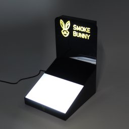 Dream Promotion - Podświetlane logo 'Smoke Bunny' na czarnym, dwupoziomowym stojaku ekspozycyjnym z podświetloną podstawą, na szarym tle.