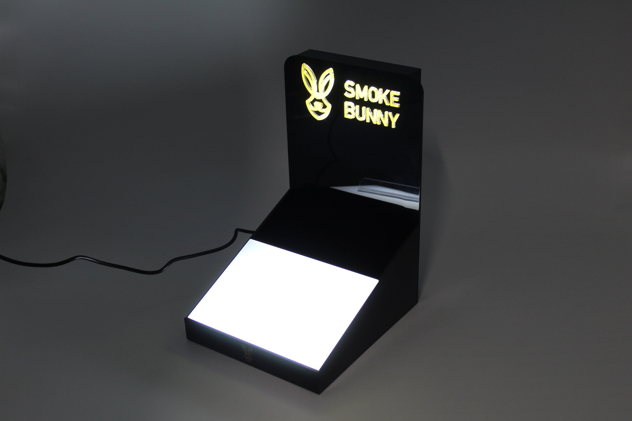 Podświetlane logo 'Smoke Bunny' na czarnym, dwupoziomowym stojaku ekspozycyjnym z podświetloną podstawą, na szarym tle.