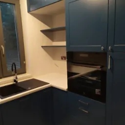 Granatowa zabudowa kuchenna z piekarnikiem w zabudowie, czarnym zlewem i białym blatem, oświetlona światłem dziennym wpadającym przez okno.