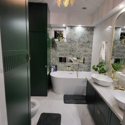 KAMILMEBLE - Nowoczesna łazienka z zabudową w kolorze butelkowej zieleni, białą wanną wolnostojącą, marmurową podłogą i złotą lampą w kształcie liści palmowych, rzucającą dekoracyjne cienie na sufit.