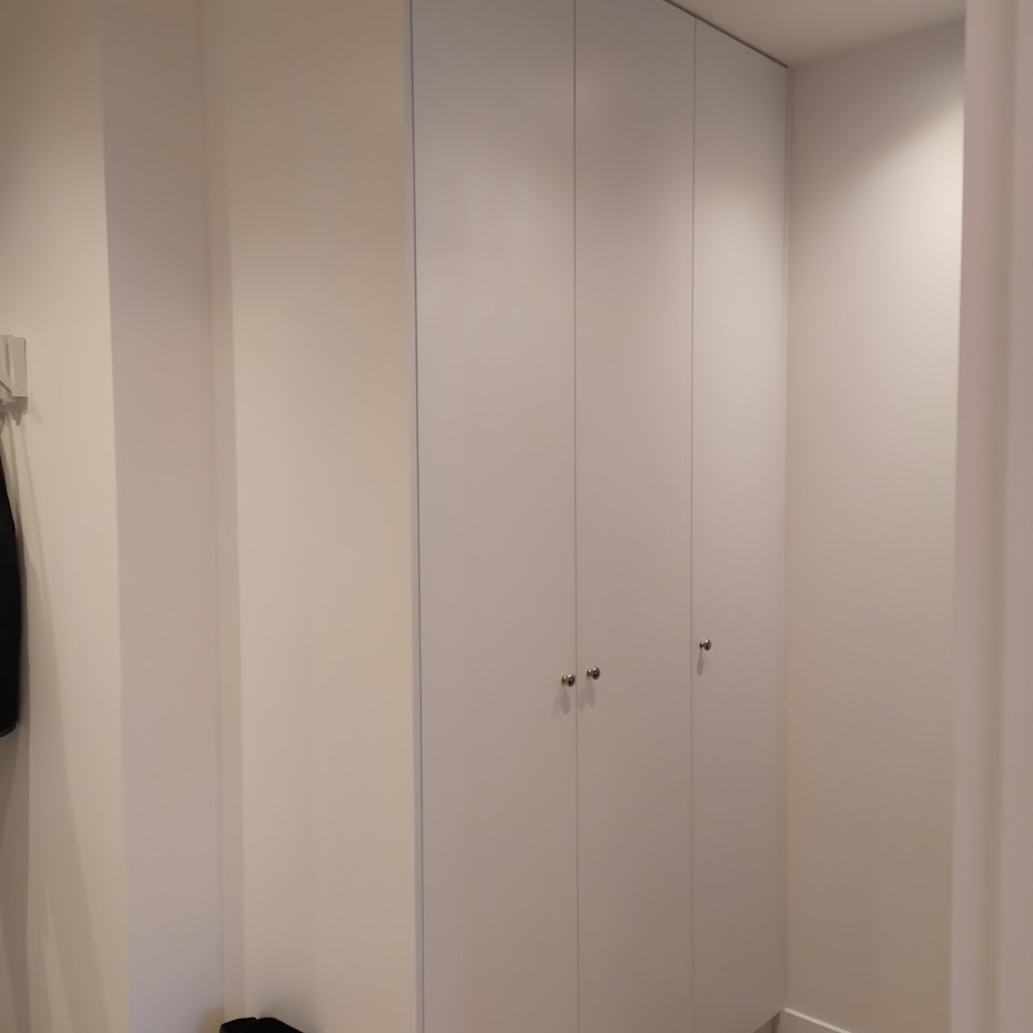 Biała, czterodrzwiowa szafa wnękowa z małymi, okrągłymi uchwytami, wkomponowana w jasne wnętrze, widoczna z perspektywy korytarza.