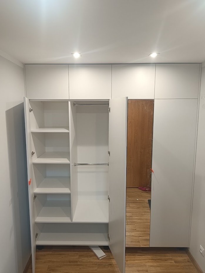 Biała zabudowa meblowa z szafą z lustrem i otwartą szafą z półkami i drążkami na ubrania, fragment listwy przypodłogowej na podłodze.