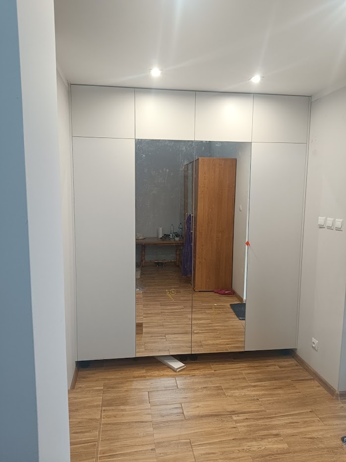 Biała szafa wnękowa z lustrzanymi drzwiami, odbijająca wnętrze pokoju z drewnianą szafą i stołem, widoczna podłoga z drewnopodobnych paneli.