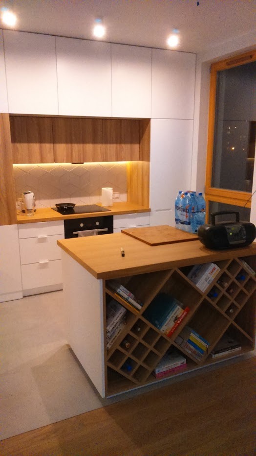 Nowoczesna kuchnia z białymi szafkami, drewnianymi akcentami i wyspą z regałem na książki i wino, oświetlenie podszafkowe LED.