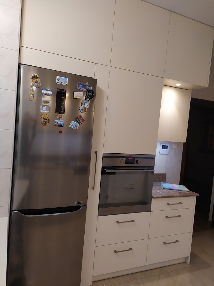 Jasna kuchnia na wymiar z zabudowaną lodówką i piekarnikiem, fronty szafek bez uchwytów, lodówka ozdobiona magnesami.