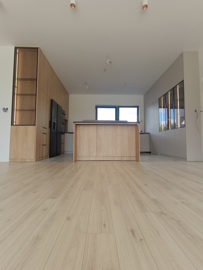 Nowoczesna kuchnia z wyspą i zabudową meblową w odcieniach jasnego drewna, minimalistyczne oświetlenie sufitowe, widok z niskiej perspektywy podkreślający przestronność wnętrza.