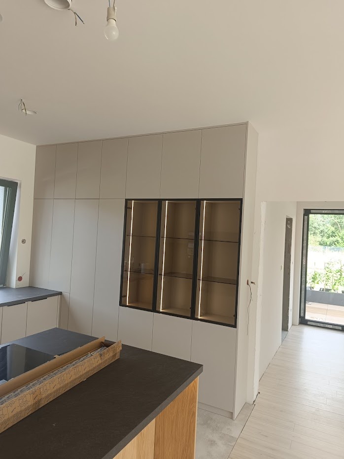 Jasna zabudowa kuchenna z wysokimi szafkami i witrynami z podświetleniem LED, widoczny fragment wyspy kuchennej z blatem w kolorze grafitu, surowy stan wykończenia wnętrza.