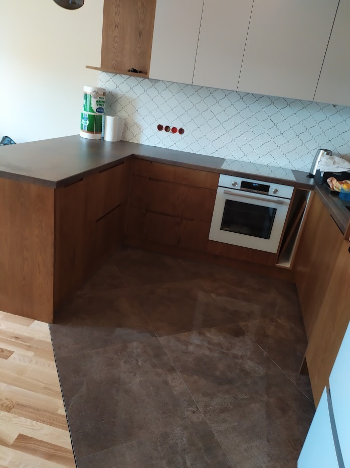 Kuchenna zabudowa narożna z drewnianymi frontami, szarym blatem i białymi szafkami wiszącymi, z piekarnikiem w zabudowie i ozdobnymi płytkami na ścianie, widoczne niedokończone otwory na gniazdka...