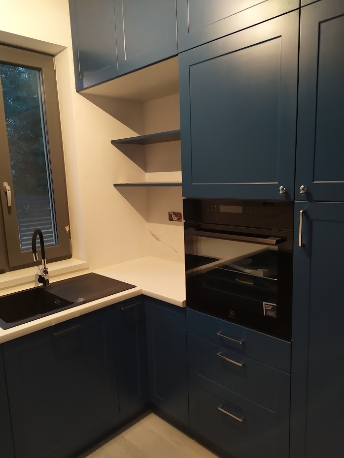 Granatowa zabudowa kuchenna z piekarnikiem w zabudowie, czarnym zlewem i białym blatem, oświetlona światłem dziennym wpadającym przez okno.