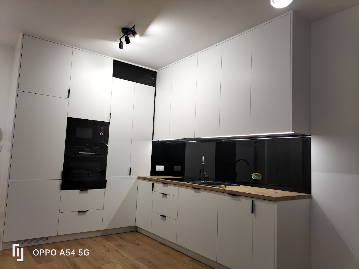 Minimalistyczna kuchnia z białymi szafkami, czarnym panelem szklanym nad blatem i drewnianym blatem, oświetlona punktowo lampami sufitowymi.