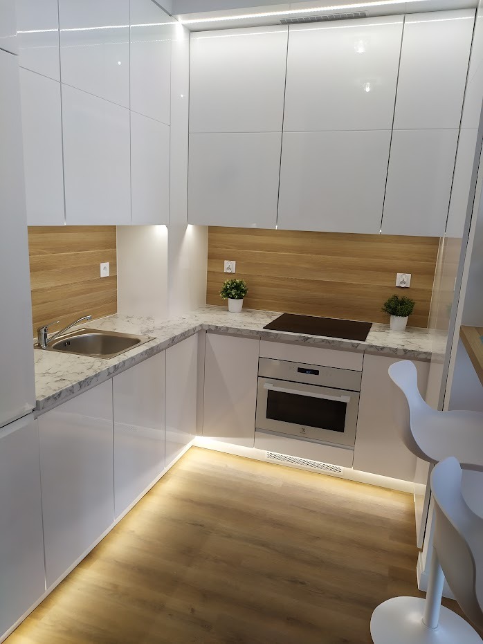 Nowoczesna, biała kuchnia z błyszczącymi frontami, blatem imitującym marmur, drewnianą ścianą nad blatem i podświetleniem LED pod szafkami.