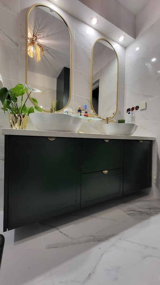 Łazienka z ciemnozieloną szafką pod umywalki nablatowe, złotymi uchwytami i owalnymi lustrami w złotych ramach na tle marmurowych płytek ściennych.