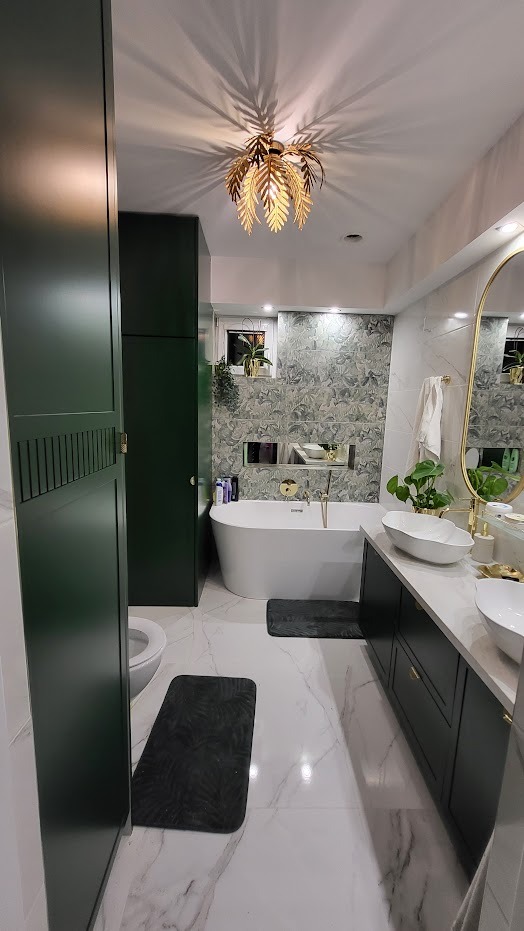 Nowoczesna łazienka z zabudową w kolorze butelkowej zieleni, białą wanną wolnostojącą, marmurową podłogą i złotą lampą w kształcie liści palmowych, rzucającą dekoracyjne cienie na sufit.