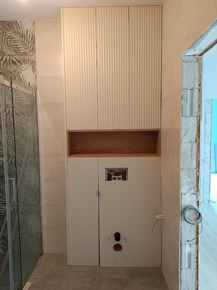 Biała zabudowa w łazience z ryflowanymi frontami szafek wiszących, drewnianą wnęką i dolną szafką z dostępem do instalacji hydraulicznej, widoczna kabina prysznicowa z wzorem liści na szkle...