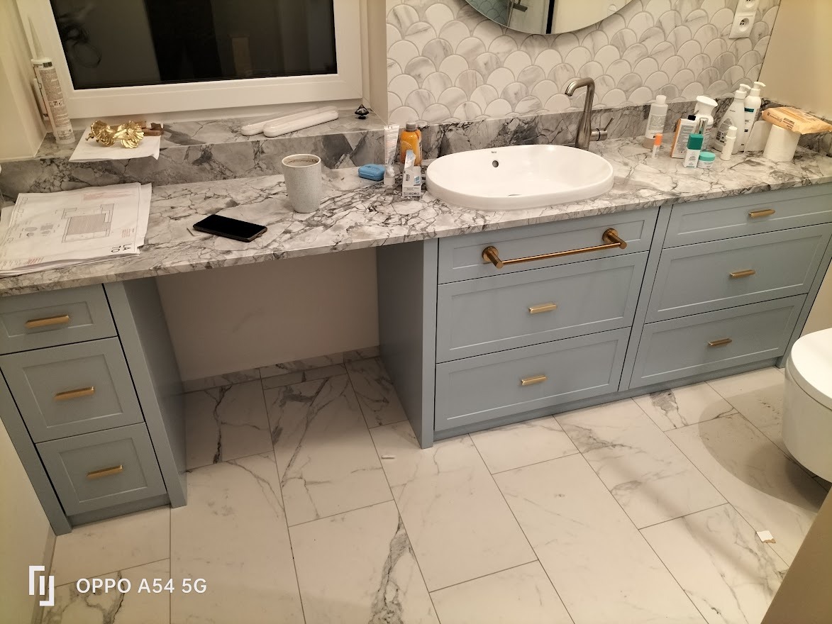 Łazienka z umywalką nablatową i szufladami w odcieniach szarości, marmurowy blat i płytki, widoczne plany na blacie, złote uchwyty.