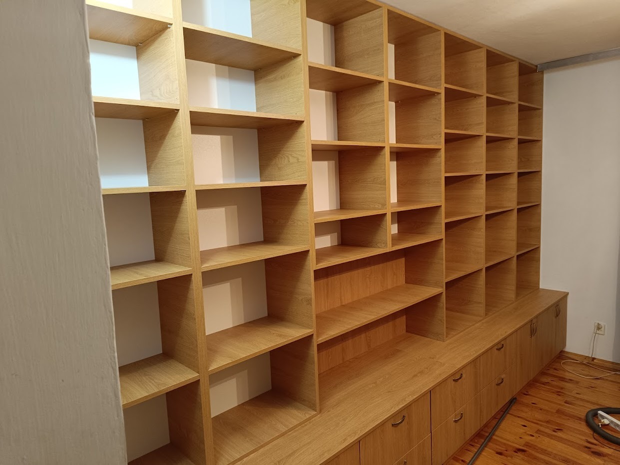 Duża, jasna, drewniana zabudowa meblowa z półkami i szafkami z szufladami, dopasowana do wymiarów pomieszczenia, z widocznymi elementami wykończenia ścian.