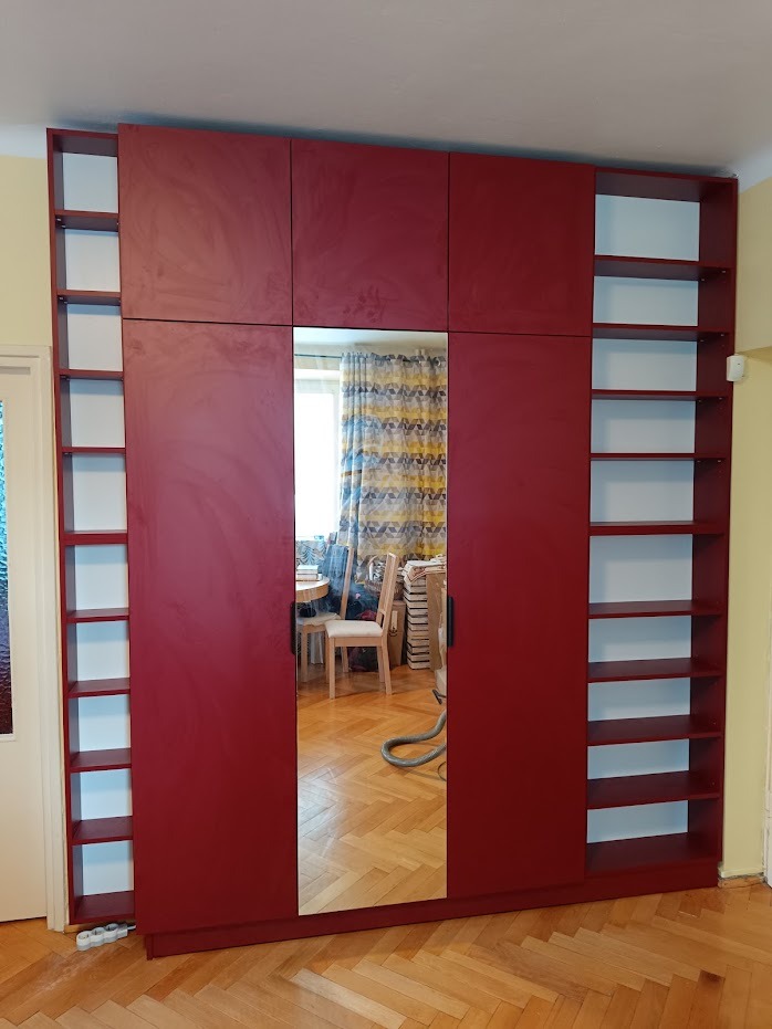 Czerwona szafa wnękowa z lustrem i półkami po bokach, widoczna w odbiciu część pokoju z krzesłem i zasłonami.