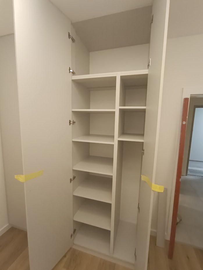 Biała, otwarta szafa wnękowa z półkami, zabezpieczona żółtą taśmą malarską, widok na wnętrze szafy i fragment pomieszczenia w trakcie remontu.