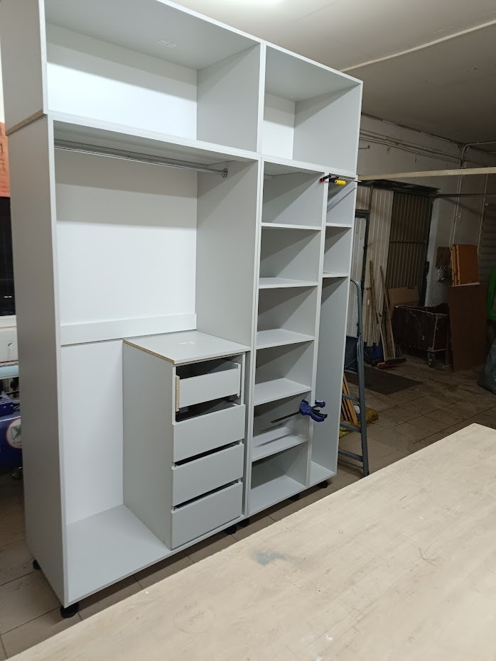 Biała, częściowo zmontowana szafa z szufladami i półkami, widoczne ściski stolarskie, pręt na ubrania i surowe wykończenie w warsztacie.