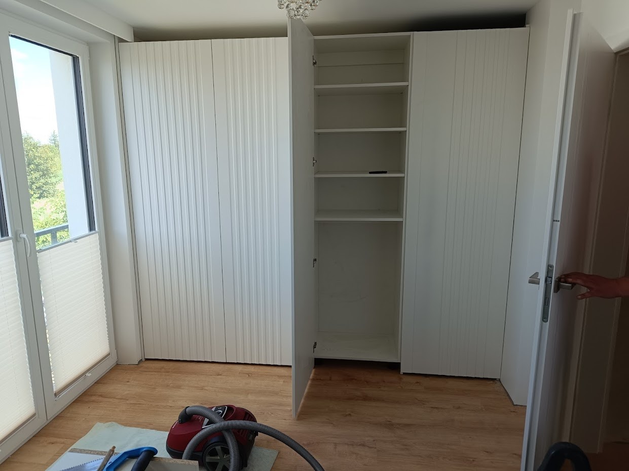 Biała szafa wnękowa z ryflowanymi drzwiami, jedna szafka otwarta ukazująca puste półki, na podłodze odkurzacz i narzędzia stolarskie.