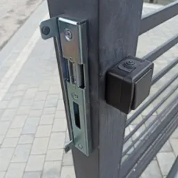 Szczegół montażu elektrozaczepu i przycisku otwierania na słupku bramy posesyjnej, widoczne śruby mocujące i fragment kostki brukowej