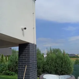 Zewnętrzna kamera monitoringu zamontowana na ścianie domu z widoczną instalacją kablową, obok jacuzzi Lay-Z-Spa St. Moritz i zielonego trawnika.