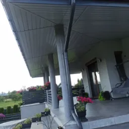 Szare orynnowanie pionowe, mocowane do kolumny podtrzymującej zadaszenie tarasu, z widocznymi elementami mocującymi, na tle kwiatów w donicach i zieleni ogrodu.