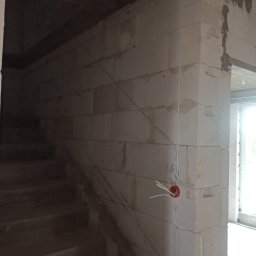 P.H . I U.E. ELBART Bartosz Lewandowski - Wewnętrzne ściany z betonu komórkowego w trakcie budowy, widoczne przewody elektryczne wystające z puszki instalacyjnej, obok schody.