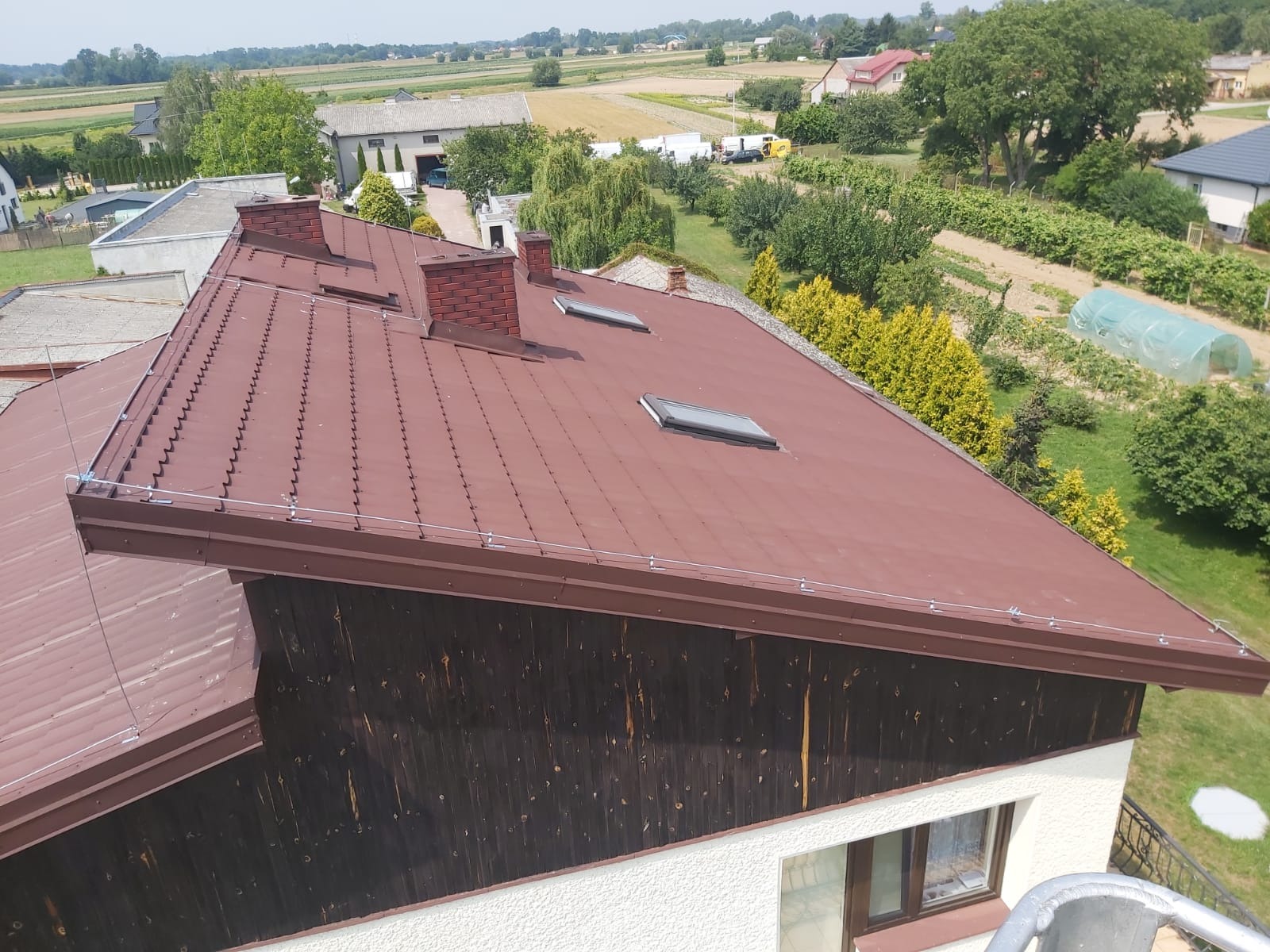 Brązowy dach z blachy trapezowej z oknami dachowymi i instalacją odgromową, widok z góry na tle wiejskiego krajobrazu.