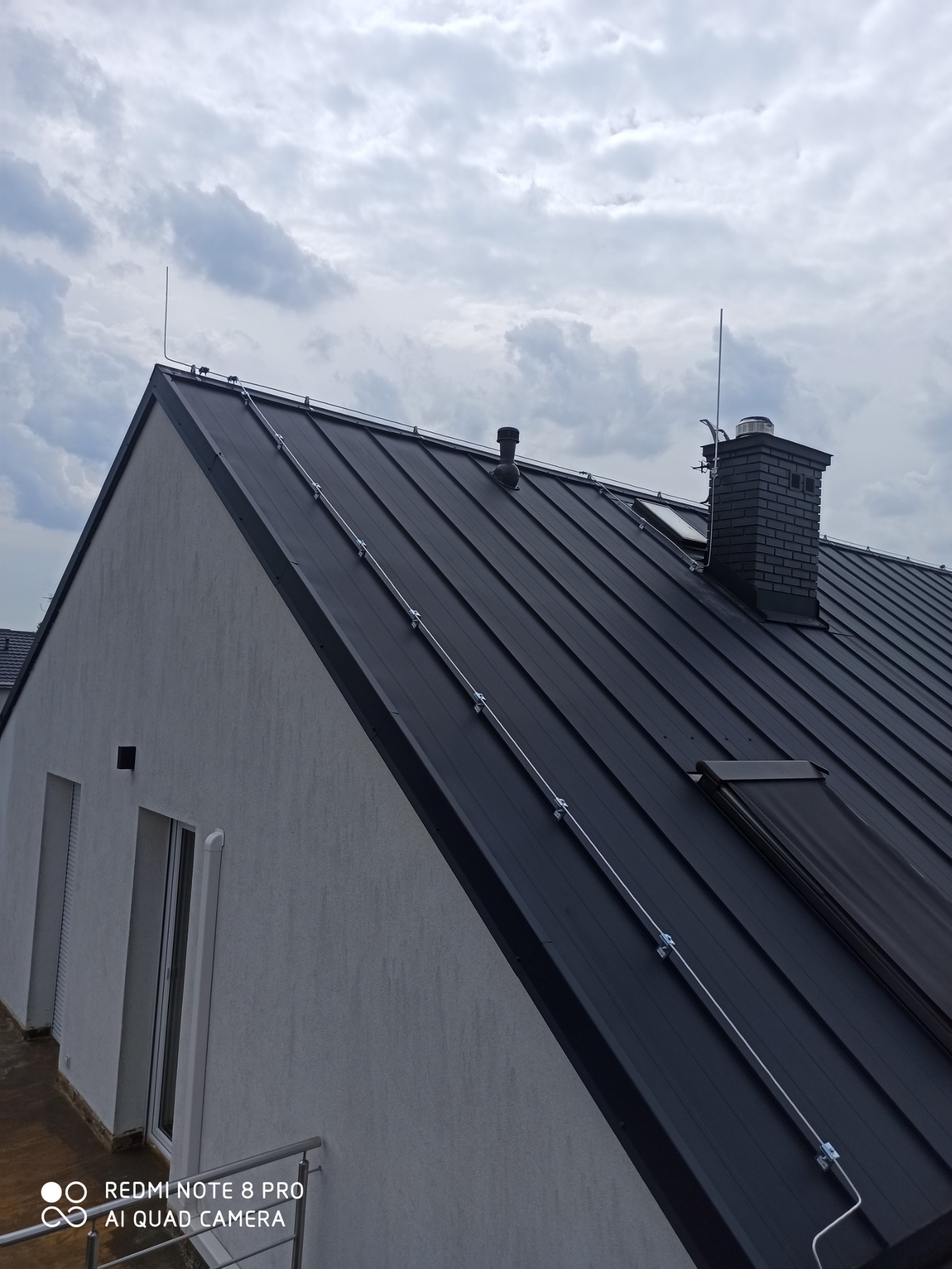 Czarny dach z blachy na nowym domu z zamontowaną instalacją odgromową, widoczne przewody i zwody pionowe, komin z cegły. Pochmurne niebo w tle.