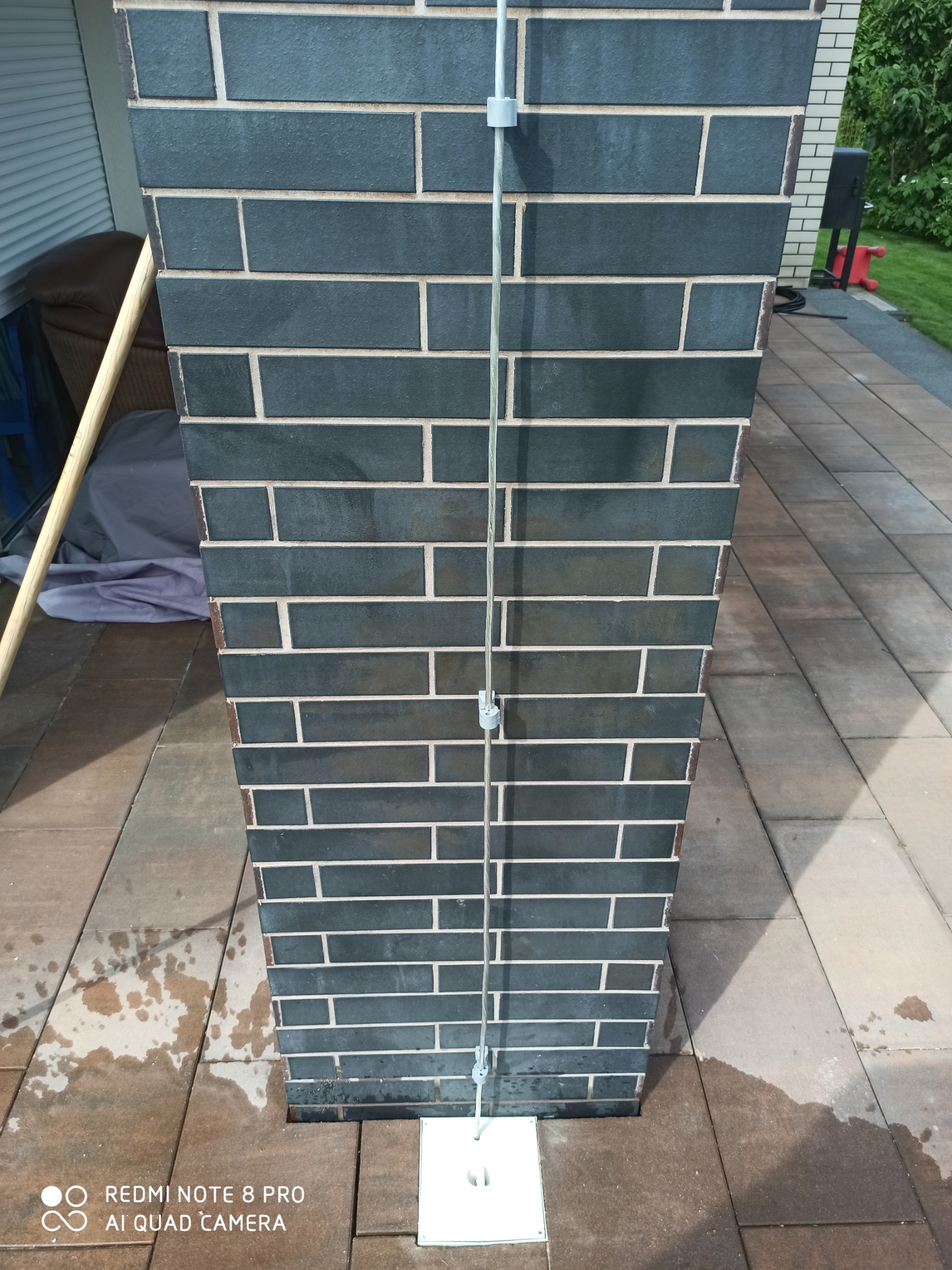 Czarny komin z cegły klinkierowej z zamontowanym przewodem odgromowym na zewnątrz budynku, widoczne mokre plamy na kostce brukowej obok.