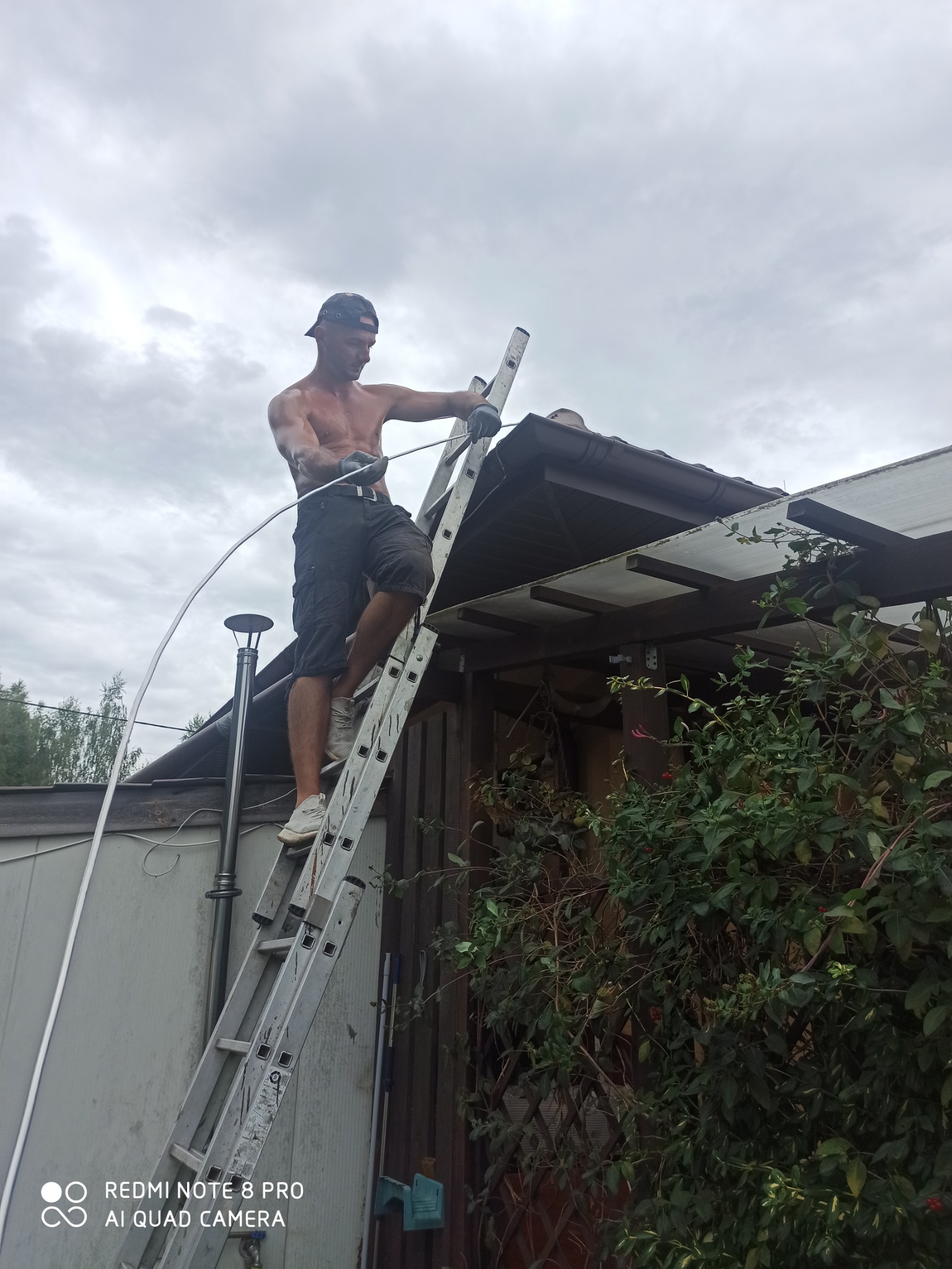 Mężczyzna bez koszulki na drabinie przy dachu, trzymający element instalacji odgromowej na tle pochmurnego nieba.