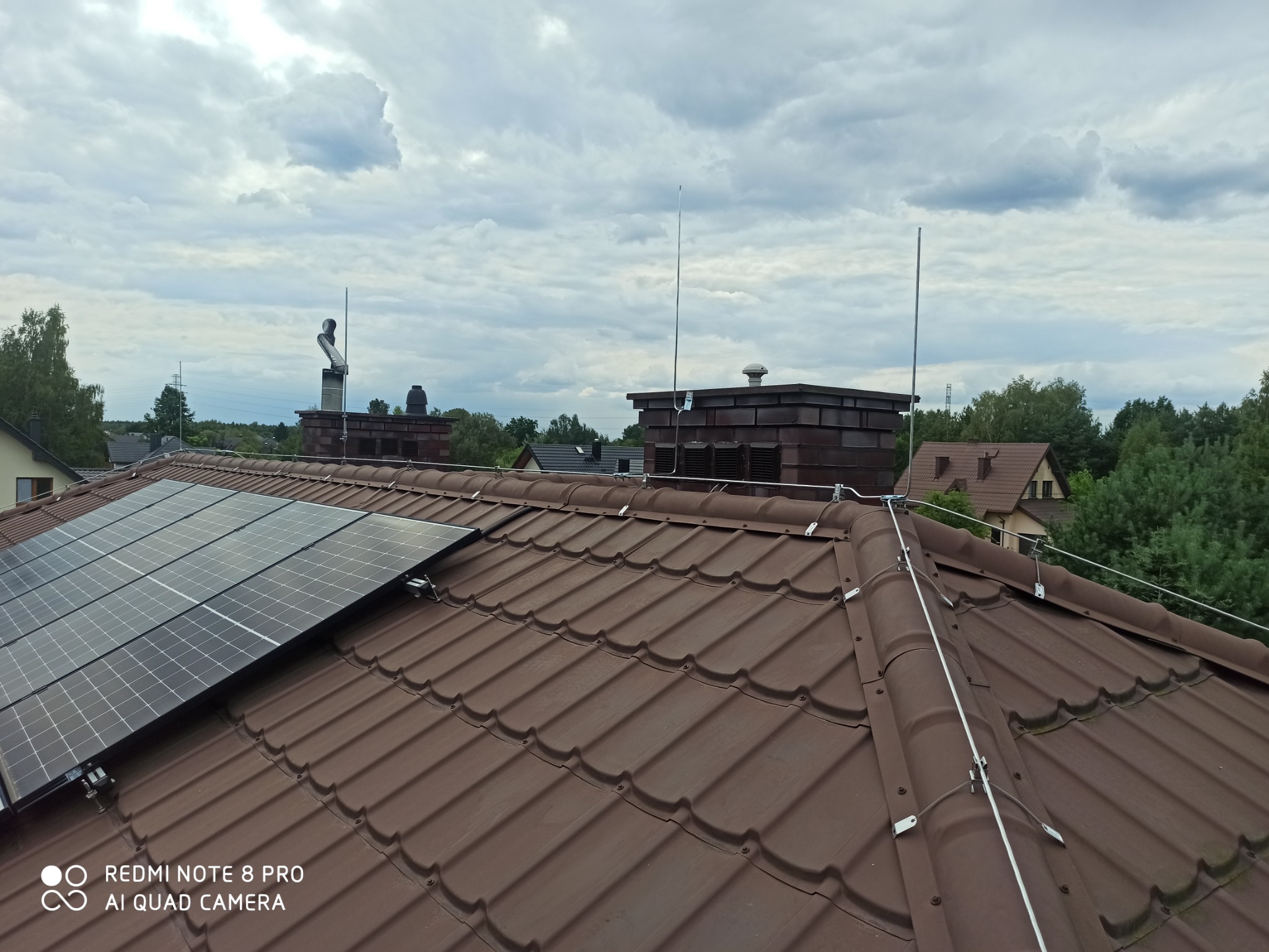 Brązowy dach z dachówki ceramicznej z zamontowaną instalacją odgromową i panelami fotowoltaicznymi, widoczne kominy i maszty odgromowe na tle pochmurnego nieba.