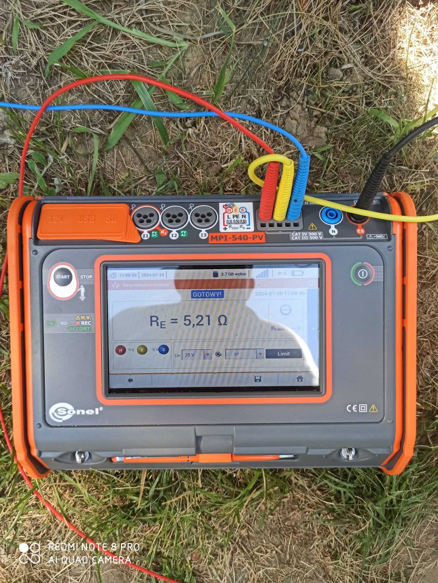 Miernik Sonel MPI-540-PV leżący na trawie, z podłączonymi przewodami pomiarowymi w kolorach czerwonym, niebieskim i żółtym, wyświetlający wynik pomiaru rezystancji uziemienia 5.21 Ω na ekranie.