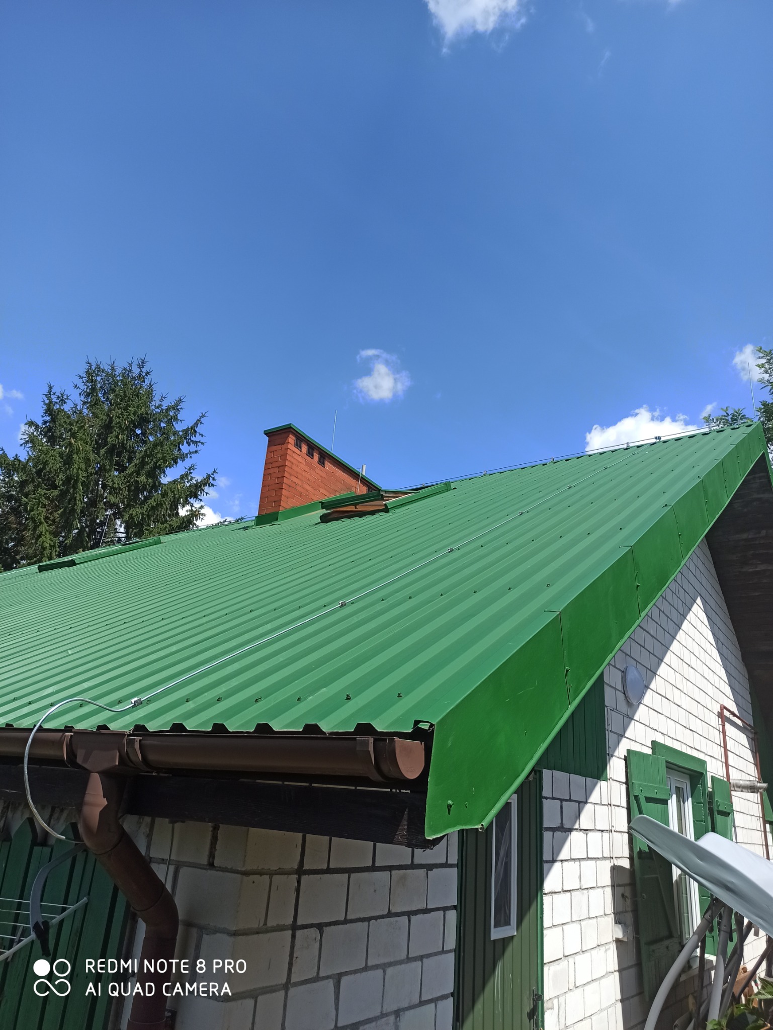 Zielony blaszany dach z brązowymi rynnami, ceglany komin z odgromnikiem na tle błękitnego nieba.