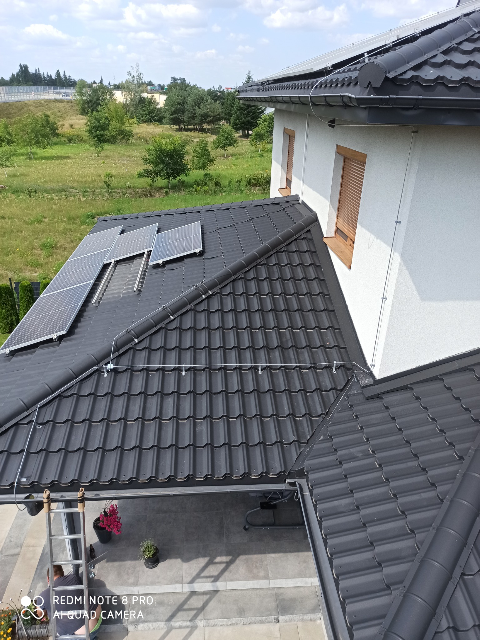 Widok z góry na czarny dach z panelami słonecznymi, widoczne przewody, rynny, drabina oparta o ścianę budynku, w tle zielony krajobraz