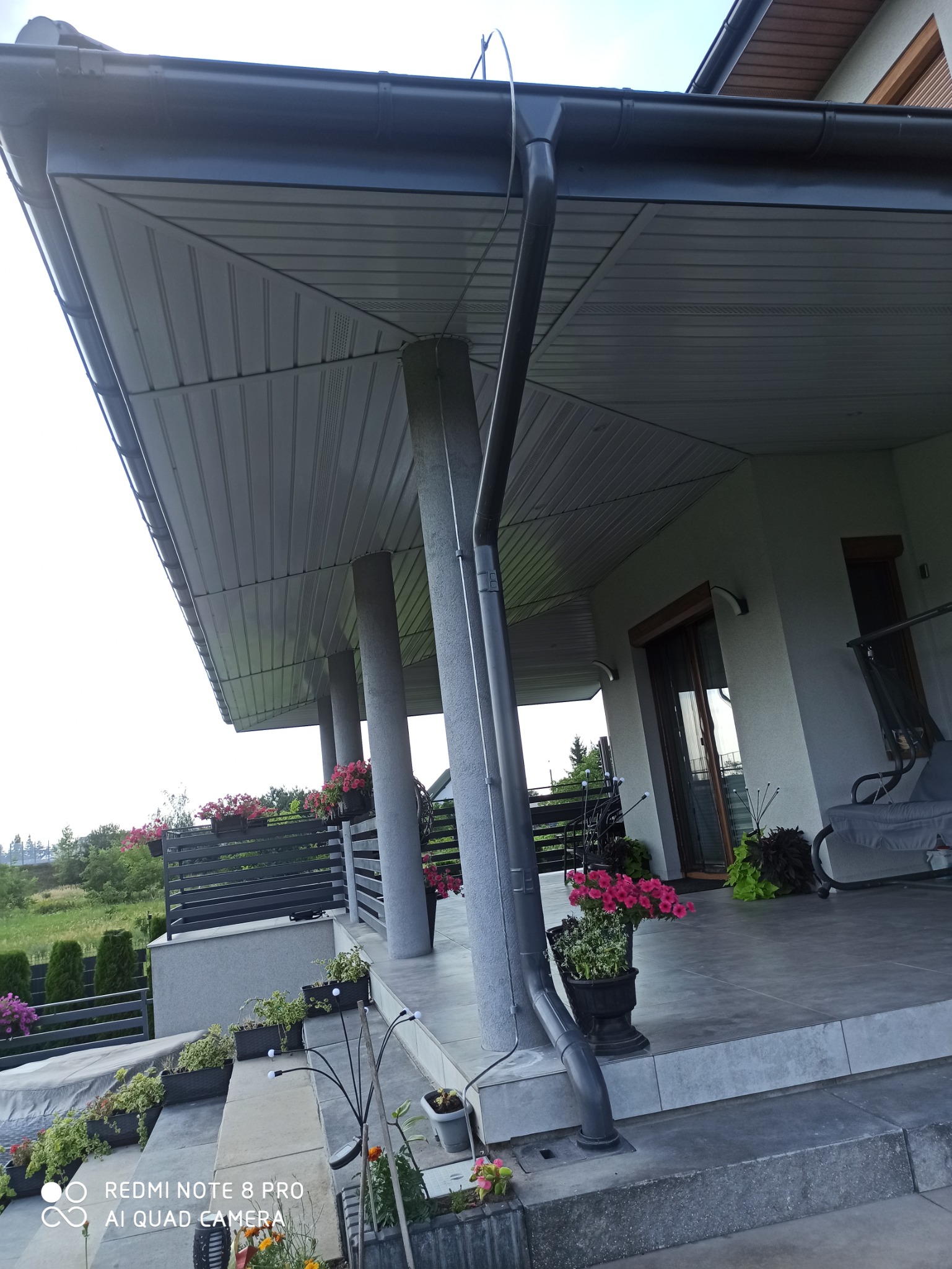 Szare orynnowanie pionowe, mocowane do kolumny podtrzymującej zadaszenie tarasu, z widocznymi elementami mocującymi, na tle kwiatów w donicach i zieleni ogrodu.
