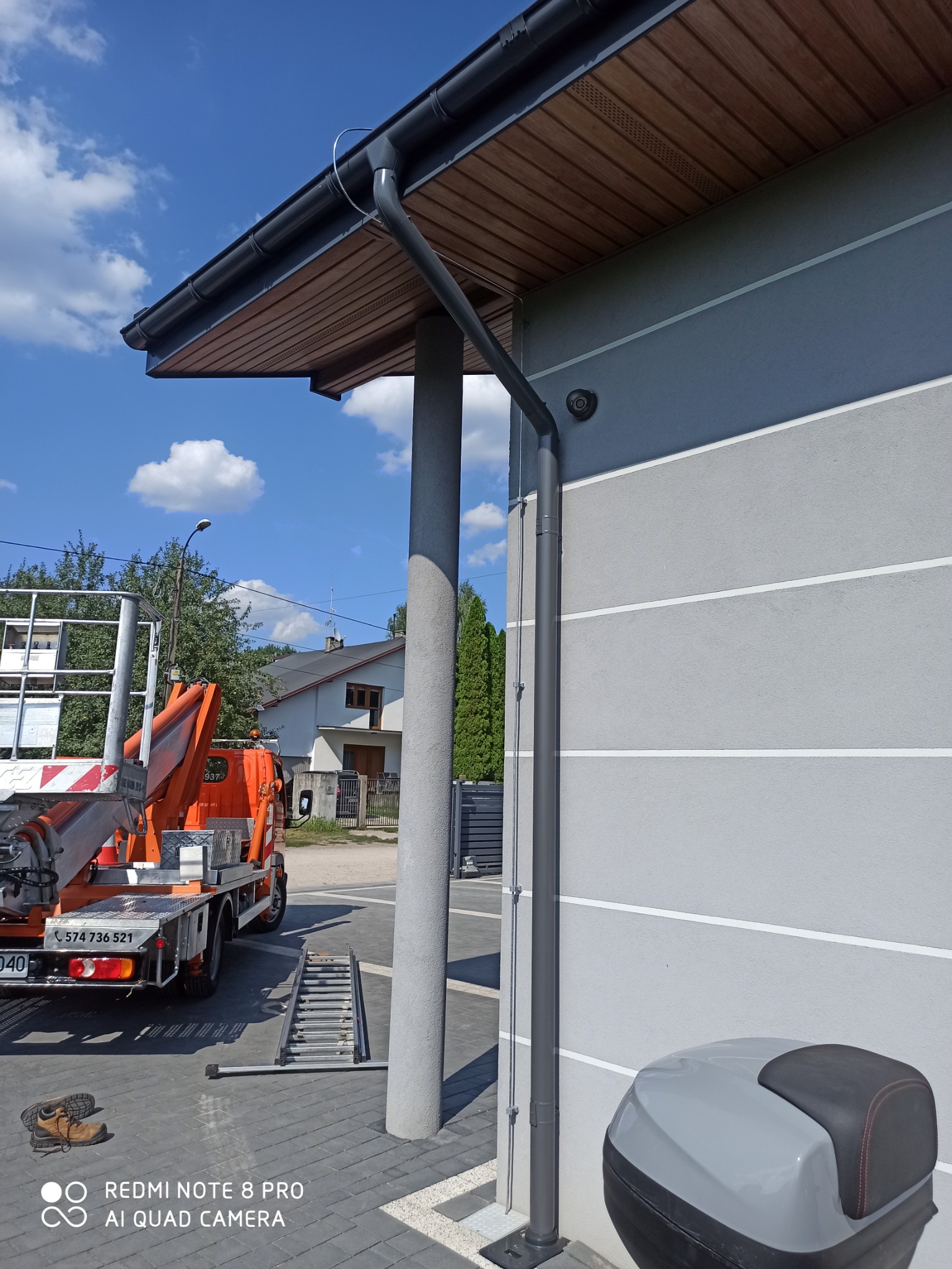 Szara elewacja budynku z drewnianą podbitką, ciemnoszara rynna spustowa i pionowa rura, na tle błękitnego nieba z chmurami, widoczny podnośnik koszowy z wysuniętą drabiną i pozostawione buty robocze.