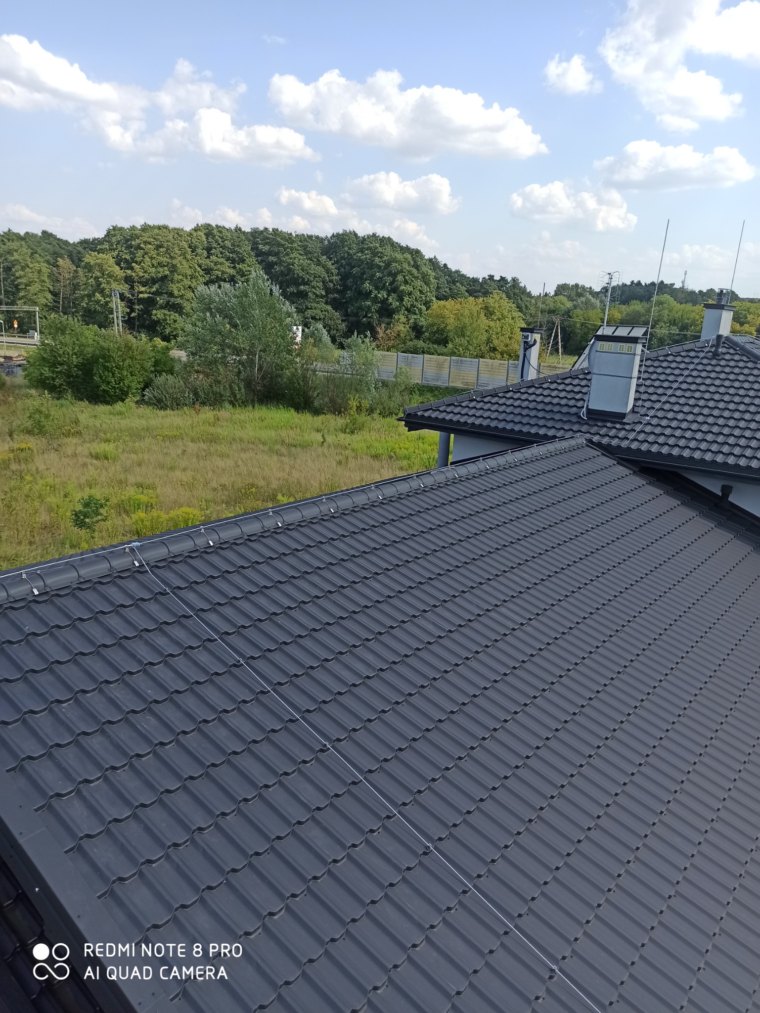 Widok z góry na dach pokryty ciemną dachówką z zainstalowaną instalacją odgromową, w tle drzewa i błękitne niebo z chmurami.