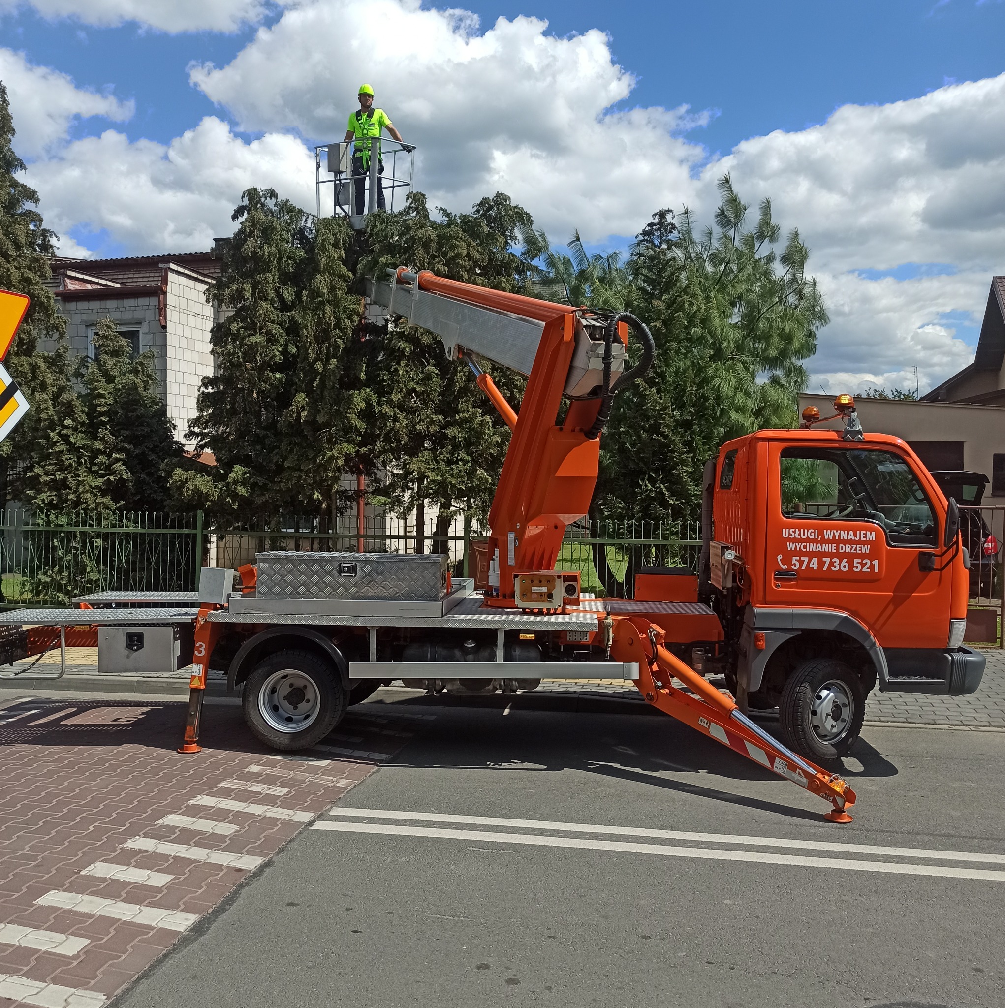Pomarańczowy podnośnik koszowy na ulicy z pracownikiem w żółtym kasku i kamizelce na platformie, w tle budynki i drzewa pod błękitnym niebem z chmurami.
