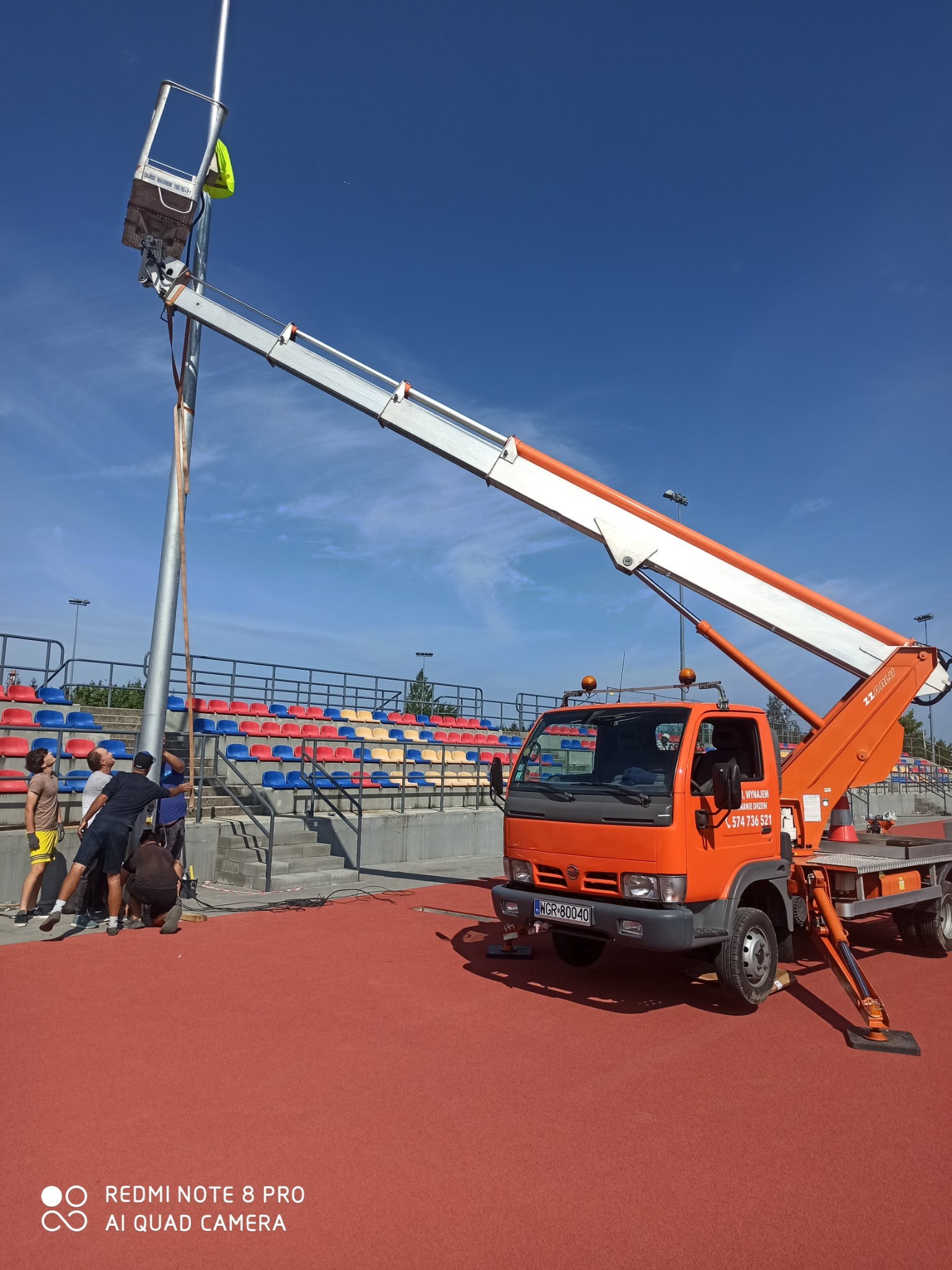 Grupa mężczyzn montuje wysoki słup oświetleniowy na stadionie, używając pomarańczowego podnośnika koszowego, na tle trybun z kolorowymi krzesełkami i błękitnego nieba.