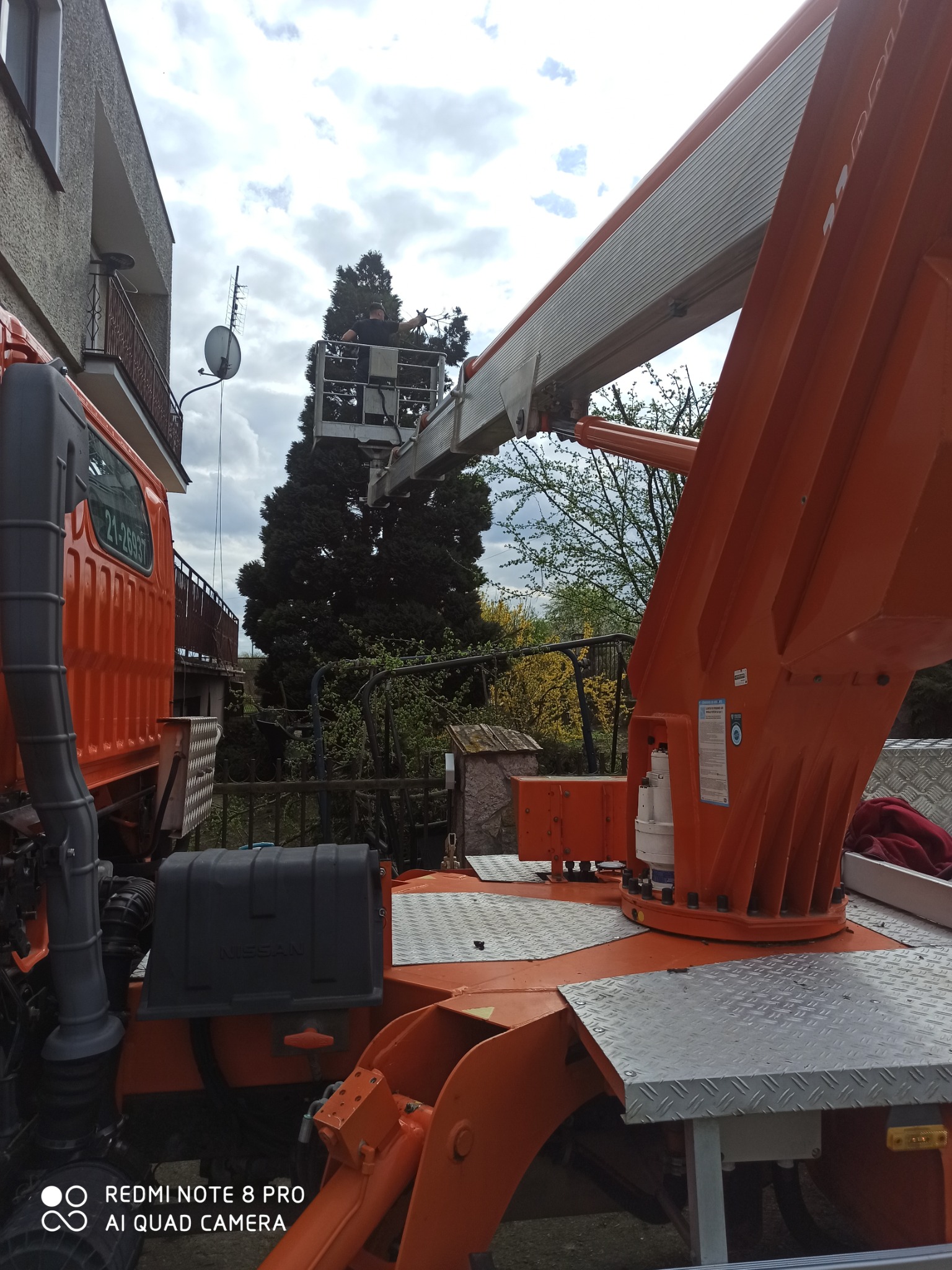 Prace elektryczne wykonywane z podnośnika koszowego przy budynku mieszkalnym, widoczna antena satelitarna na balkonie.