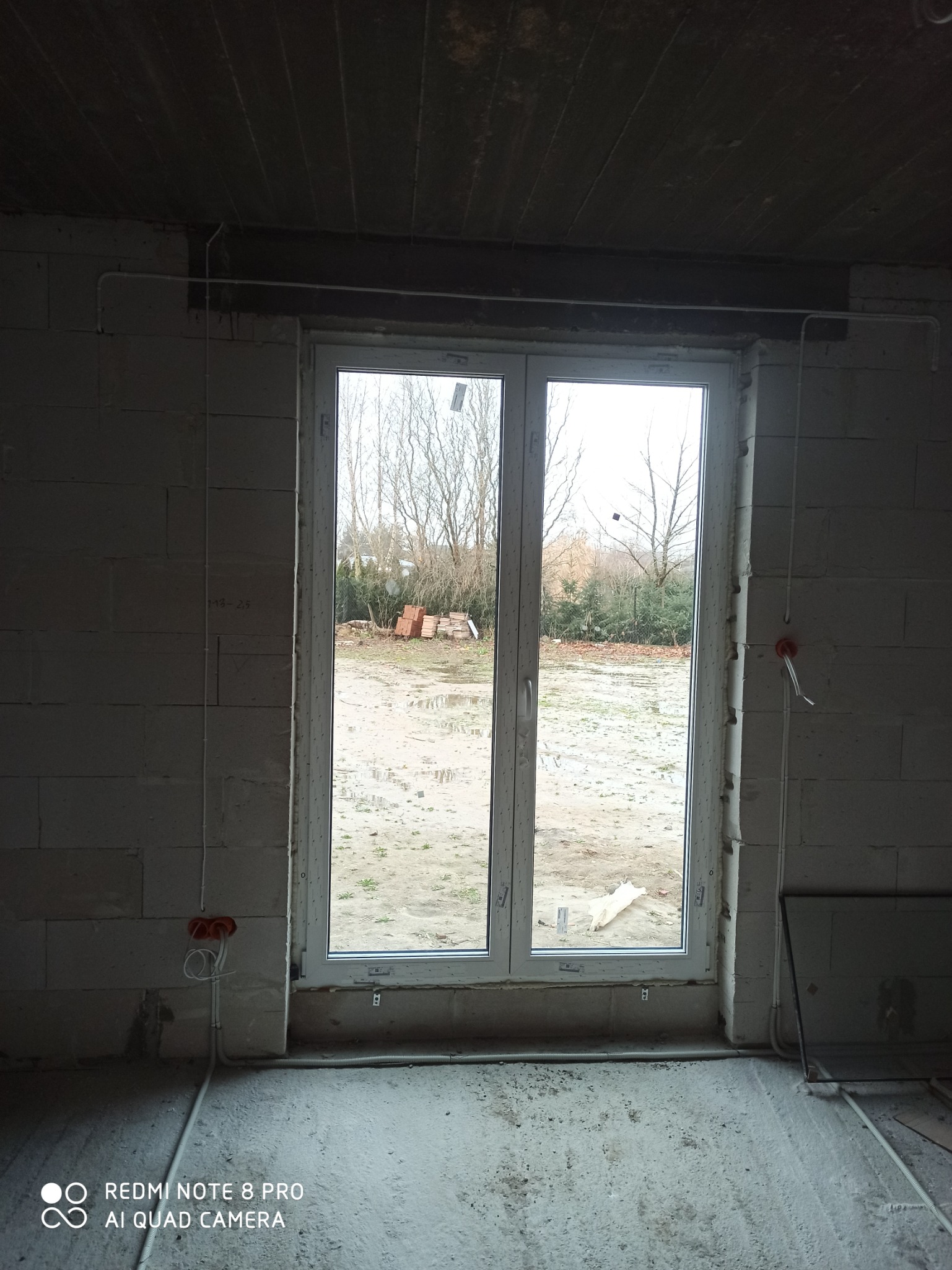 Wnętrze surowego budynku z oknem, widoczne rozprowadzone przewody elektryczne w peszlach i puszki instalacyjne w ścianach z betonu komórkowego.