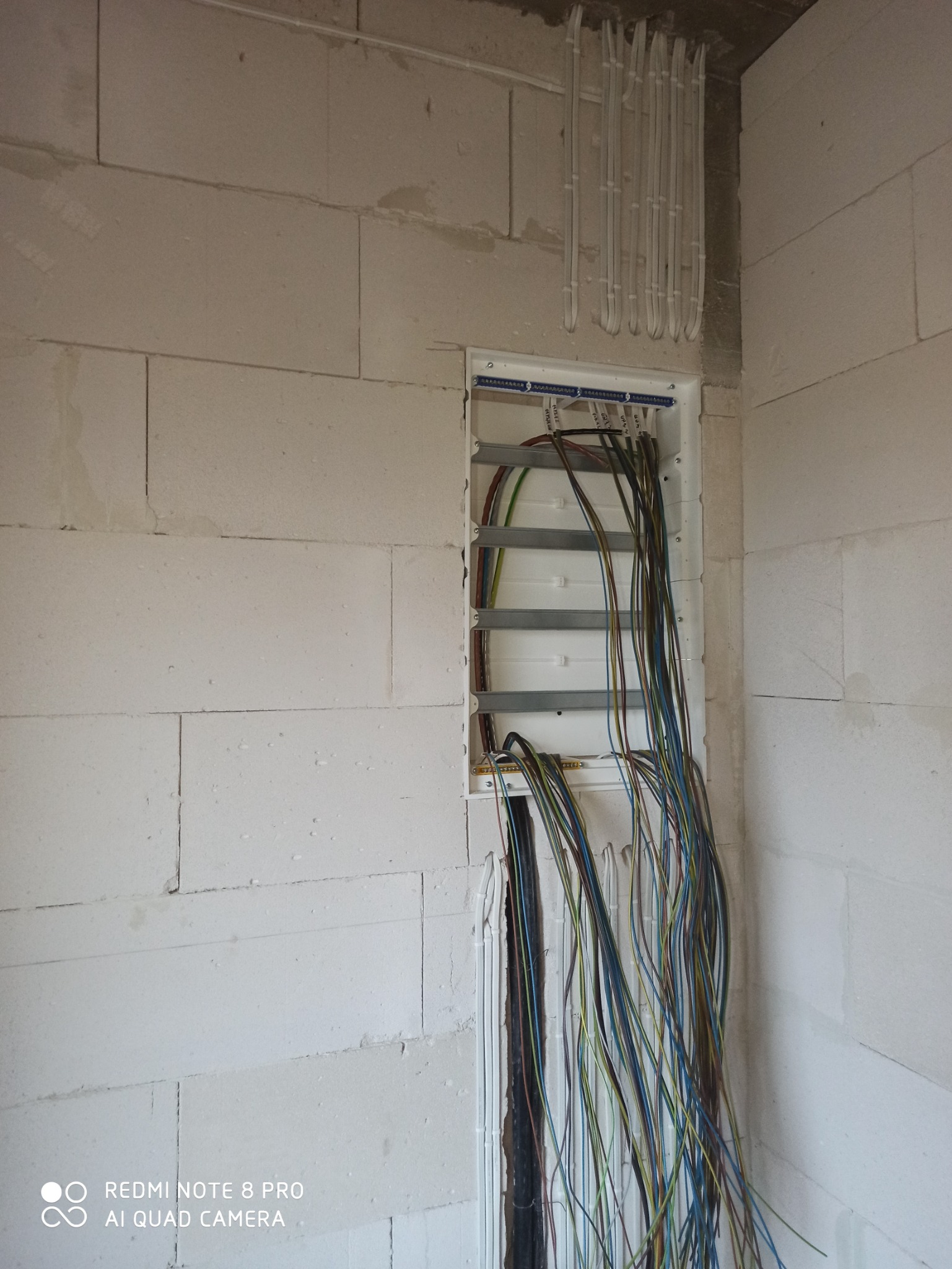 Rozdzielnica elektryczna w trakcie montażu w surowym wnętrzu, widoczne przewody w różnych kolorach, kable prowadzone w peszlach na ścianie z białych bloczków.