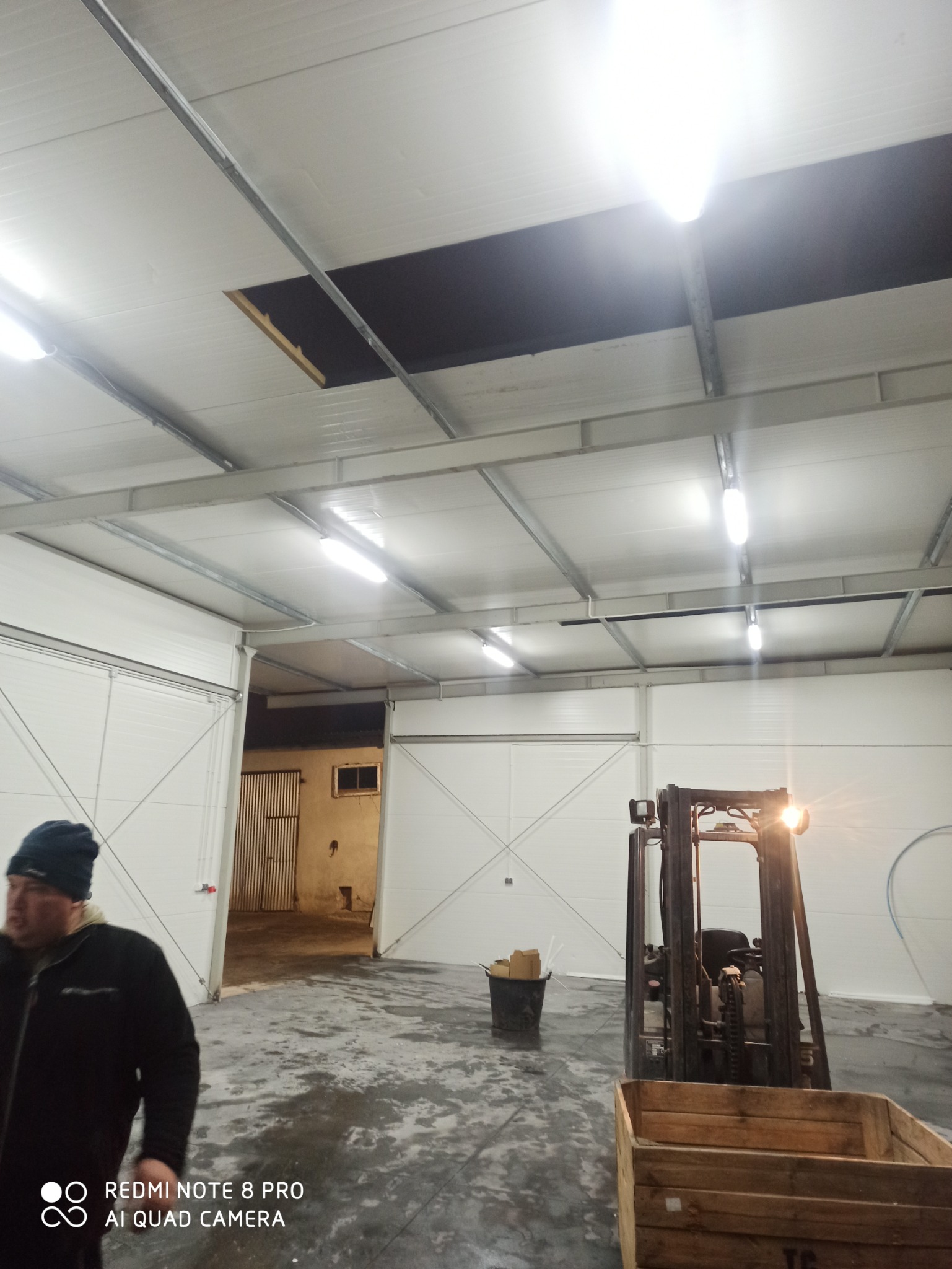 Wnętrze hali magazynowej z widocznym wózkiem widłowym, otwartą bramą i niedokończonym sufitem podwieszanym z zamontowanym oświetleniem LED. Fragment sylwetki mężczyzny w lewym dolnym rogu.