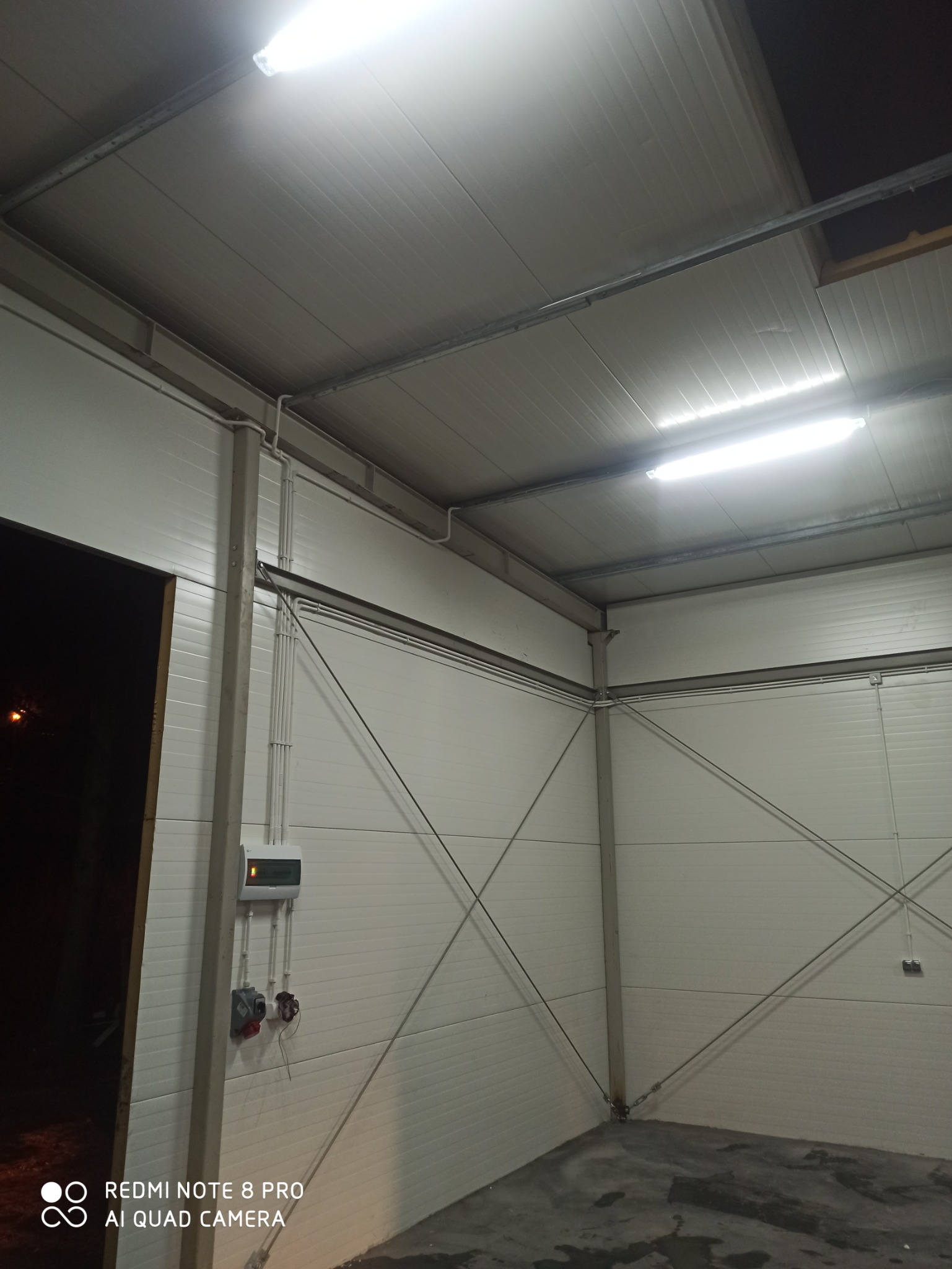 Wnętrze hali produkcyjnej z widoczną instalacją elektryczną: kable w peszlach, skrzynka rozdzielcza, gniazda siłowe i oświetlenie jarzeniowe pod sufitem.